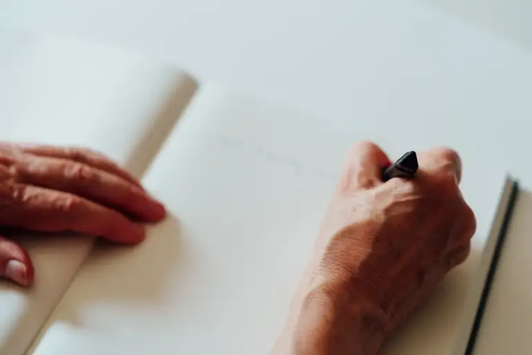 Ein aufgeschlagenes Buch, die linke Hand ruht auf der geöffneten Buchseite, die rechte Hand hält einen schwarzes Stift und schreibt.
