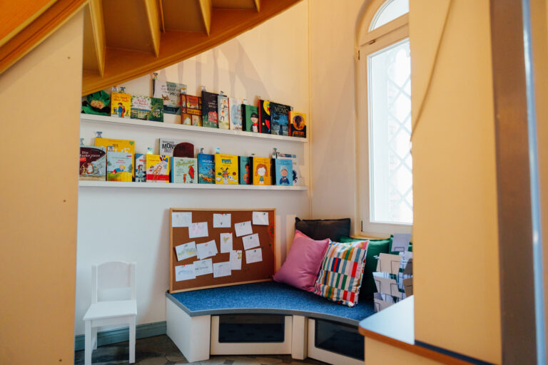 Unter einer Wendeltreppe ist eine gemütliche, mit Sitzkissen ausgestattete Leseecke. An der Wand sind auf einer Bilderleiste reichlich Kinderbücher aufgereiht.