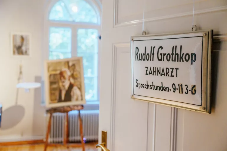 Emailliertes Schild hängt an einer geöffneten Tür mit der Aufschrift: "Rudolf Grothkop, Zahnarzt, Sprechstunden 9-11, 3-6". Im Hintergrund ist eine Staffelei mit Leinwand zu sehen.