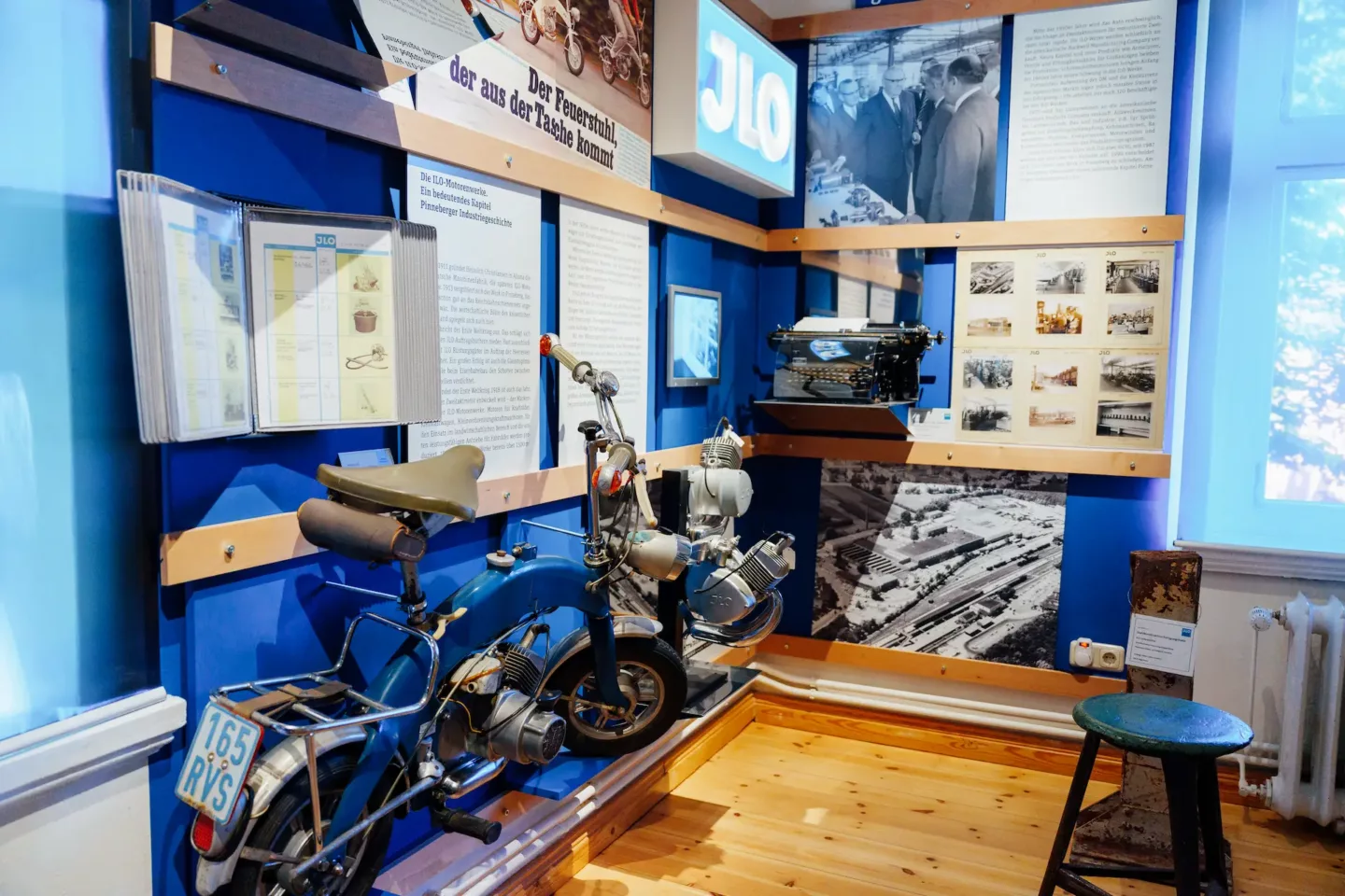 Ein Ausstellungsraum, durchzogen von der Farbe blau, zeigt Motor, Schreibmaschine, Leuchtreklame und Klappmotorad der ILO Motorenwerke.