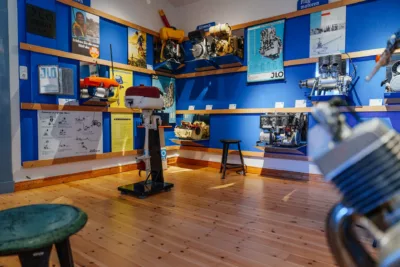 Diverse Motoren füllen den Ausstellungsraum: für Boote, Schneeschlitten und Helikopter sowie begleitende Werbematerialien und technische Zeichnungen sind an und vor einer prägnant blauen Wand im Ausstellungsraum zu sehen.