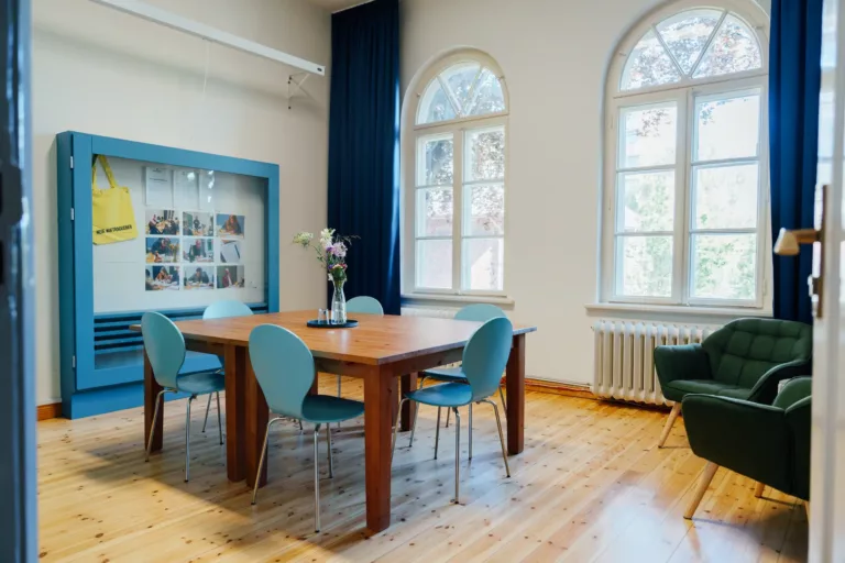 Ein heller einladender Raum mit zwei großen Fenstern, einem großen Tisch, bestuhlt, sowie weiteren Sesseln als Sitzmöglichkeit. Ein großer Schaukasten an der Wand zeigt aktuelle Projekte.